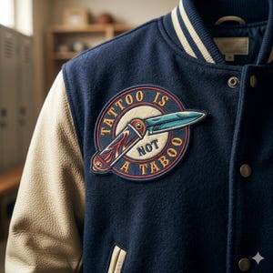 May include: A navy blue varsity jacket with cream-colored sleeves and a circular embroidered patch on the chest. The patch features a folding pocket knife graphic and the text "TATTOO IS NOT A TABOO" in gold lettering against a dark blue background.