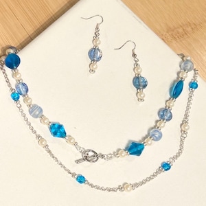 May include: A jewelry set consisting of a necklace and a pair of drop earrings. The necklace features a silver-tone chain with a toggle clasp, accented by blue glass beads, white faux pearls, and small silver spacers. The earrings match the necklace.