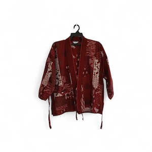 May include: A burgundy kimono-style jacket with a geometric pattern in off-white. The jacket has three-quarter sleeves and a contrasting red trim along the edges. It hangs on a black hanger against a white background.