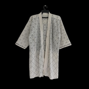 May include: White kimono-style robe with a geometric black dotted pattern. The robe has a relaxed fit with wide sleeves and a contrasting cream-colored trim along the edges and lapels. The robe is hanging on a black hanger against a black background.