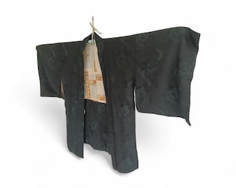 May include: A dark kimono with a floral pattern hangs against a white background. The kimono is black with a subtle design, and the interior lining is a cream color with a geometric pattern. The garment is suspended by a natural-colored cord.