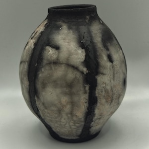 Puede incluir: Un jarrón de cerámica redondo con cuello estrecho y superficie texturizada. El recipiente presenta un patrón moteado en tonos negro mate y blanco roto, con bandas verticales negras a los lados y un borde y base de color negro sólido.