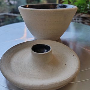 May include: A handmade ceramic bowl and lid set. The bowl is a light beige color with a dark rim. The lid is a matching beige with a small, dark-rimmed cylinder in the center. The set has a rustic, textured appearance.