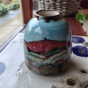 May include: A handmade ceramic vase with a unique landscape design. The vase features a blue sky, a red and brown mountain range, and earth-toned layers. The vase has a glossy finish and a dark brown rim.