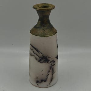 May include: A cylindrical ceramic vase with a narrow neck and a flared rim. The body of the vase is matte white with dark, smoky black marbling patterns. The neck and rim feature a dark, metallic bronze-toned finish with a textured appearance.
