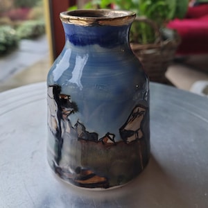 Op de afbeelding: Handgemaakte keramische vaas met een blauwe en gouden rand. De vaas heeft een landschapsontwerp met tinten blauw, bruin en wit. De vaas is een uniek kunstwerk, perfect voor het tentoonstellen van bloemen of als decoratief item.