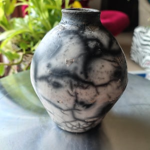 May include: A handcrafted ceramic vase with a distinctive, textured finish. The vase displays a black and white marbled pattern with a dark rim. The rounded body narrows to a slender neck, exhibiting a rustic appearance.
