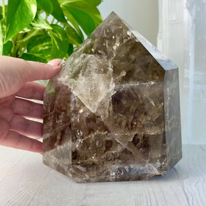 May include: A large, polished smoky quartz crystal tower with a hexagonal base and a pointed top. The crystal features a translucent, deep brown color with visible internal fractures, natural inclusions, and varying levels of clarity throughout.