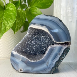 May include: A polished, blue-grey agate geode with a sparkling crystal interior. The geode has bands of color and a natural, organic shape. The geode is a decorative item, perfect for home decor or as a unique gift.