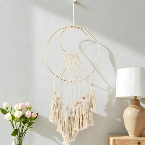 以下が含まれることがあります： 金色の金属とクリーム色のマクラメの壁掛け。デザインは、交差する2つの円、クリーム色のタッセル、木製のビーズが特徴です。壁掛けは白いコードから吊り下げられています。