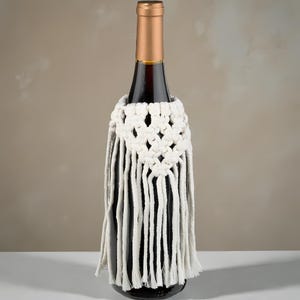Peut inclure: Une bouteille de vin sombre avec un bouchon doré est ornée d'une housse de bouteille de vin en macramé blanc. La housse présente un motif géométrique en haut et de longues franges.
