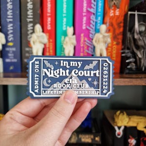 Könnte beinhalten: Ein blaues und silbernes Ticket mit dem Text "In my Night Court era Book Club" und "Lifetime Membership". Das Ticket hat ein Berg- und Fledermaus-Design. Das Ticket wird vor einem Bücherregal mit Büchern gehalten.
