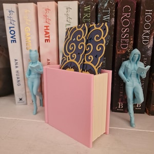 May include: A light pink book-shaped holder containing two dark blue rectangular cards with gold cloud patterns. The holder is flanked by two light blue figurines, one depicting a person with a helmet and one depicting a person in a hoodie.