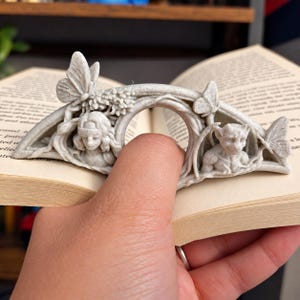 Könnte beinhalten: Ein hellgrauer, gesprenkelter Buchseitenhalter aus Kunstharz mit bogenförmigem Design, der links eine menschliche Figur und rechts eine Wasserspeier-Figur zeigt. Das Stück ist mit dreidimensionalen Schmetterlingsakzenten und Blattdetails verziert.