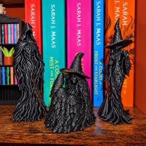 May include: Three black figurines standing on a wooden surface. The left figure is a hooded entity. The centre figure is a seated person wearing a pointed hat with a bird perched on their arm. The right figure is a standing person with a pointed hat holding a broom.