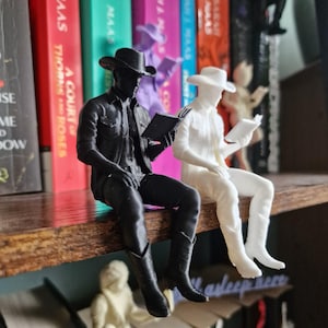 May include: Two cowboy figurines are sitting on a shelf edge. One figurine is solid black and the other is solid white. Both figures wear cowboy hats and boots, and each holds an open book in their hands, appearing to be reading while seated.