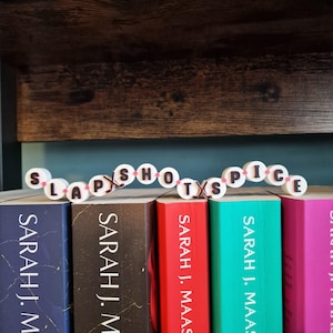 Può includere: Una catena decorativa di perline circolari bianche con lettere nere che compongono la scritta SLAP SHOTS AND SPICE, separata da piccole icone di bastoni da hockey marroni, appoggiata orizzontalmente sui bordi superiori di cinque libri.