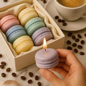 Puede incluir: Una vela de macaron lavanda encendida sostenida en una mano, con una caja de velas de macaron de colores pastel en el fondo. También se ve una taza de café y granos de café.