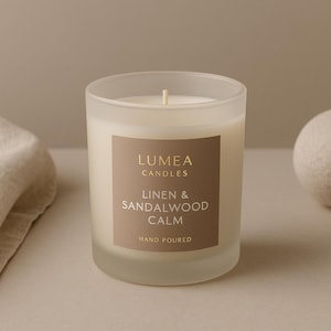 Könnte beinhalten: Eine Kerze aus mattiertem Glas mit cremefarbenem Wachs und einem Etikett mit der Aufschrift „Lumea Candles Linen & Sandalwood Calm Hand Poured“. Ein beiges Leinentuch und eine beige Kugel sind ebenfalls im Bild.