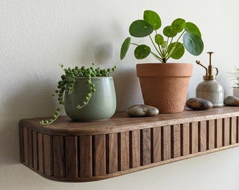 May include: A wooden floating shelf with a ribbed front holds various potted plants and decorative items. A green plant in a ceramic pot, a terracotta pot with a round-leaf plant, a small spray bottle, and a white pot with a spiky plant are displayed.