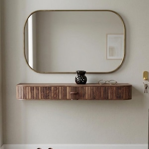 Op de afbeelding: Een moderne entree met een rechthoekige spiegel met gouden frame, een houten console met een lade en een zwarte vaas. Een bruine paraplu staat in een metalen houder. Bruine leren schoenen op een grijze mat.