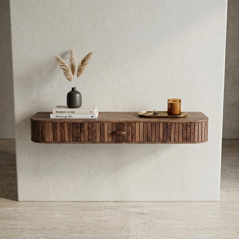 May include: A floating wooden shelf with a drawer, featuring a ribbed front design. The shelf holds a small vase with dried plants, two books, a candle, and a small tray. The books are titled "Modern Living" and "Kintsugi Art".