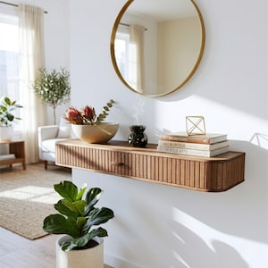 May include: A wooden floating shelf with a drawer, holding a decorative bowl with flowers, books, and a black essential oil diffuser. A gold-framed round mirror hangs above. A potted plant sits below.