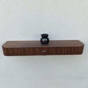 以下が含まれることがあります： ダークブラウンの木製フローティングシェルフ。引き出しと垂直の溝のディテールがあります。白い花の模様の小さな黒い装飾品が上にあります。シェルフは白い壁に取り付けられています。