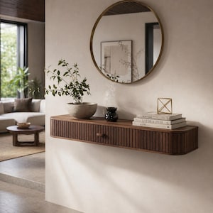 Op de afbeelding: Een zwevend plankje van donker hout met een lade, met een kleine plant in een stenen pot, boeken en een decoratief goudkleurig object. Boven het plankje hangt een ronde spiegel. Het plankje heeft een geribbeld ontwerp.