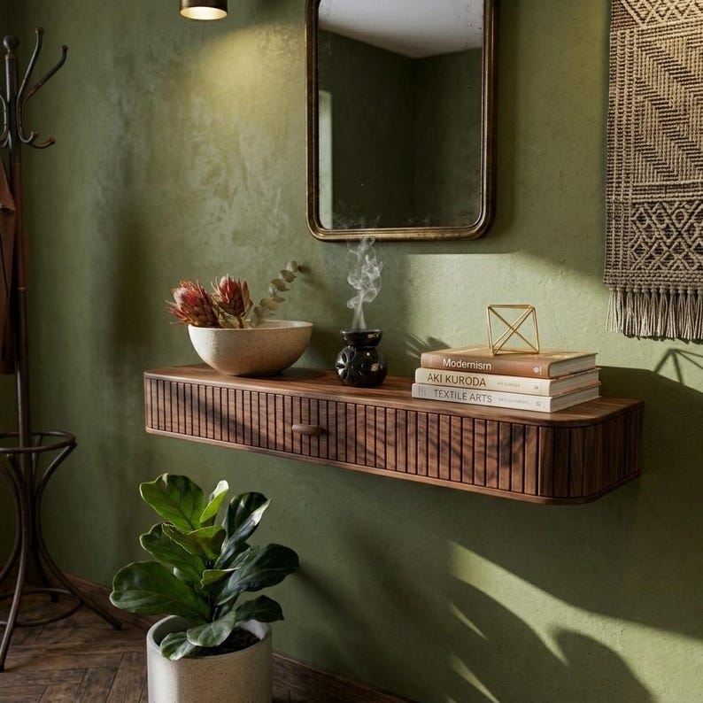 May include: A wooden floating shelf with a drawer, holding a bowl of flowers, books, and a small black incense burner. A mirror hangs above the shelf, and a potted plant sits below. The wall is painted a muted green.