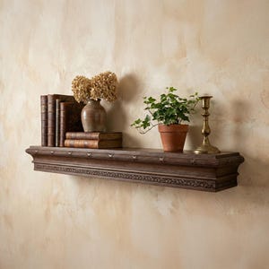 May include: A dark brown, carved wooden shelf fixed to a wall. It holds a display of old books, a vase with dried flowers, a potted plant, and a brass candlestick. The wall has a textured, beige finish, creating a classic, decorative display.
