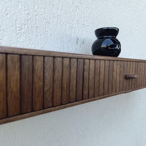 以下が含まれることがあります： ダークブラウンの木製フローティングシェルフ。引き出しと小さな黒い装飾花瓶が付いています。シェルフは垂直のリブデザインで、小さく丸みを帯びたハンドルが付いています。花瓶は光沢があり、花の模様があります。