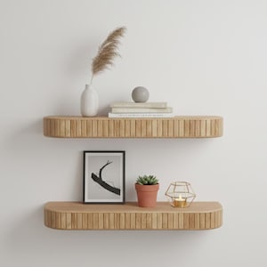 Puede incluir: Dos estantes flotantes de madera con un diseño acanalado están montados en una pared blanca. El estante superior contiene un jarrón blanco con hierba seca, una esfera gris y libros. El estante inferior muestra una impresión enmarcada, una suculenta en maceta y un candelabro.