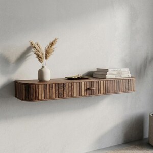 May include: A floating wooden shelf with a drawer and a curved front. The shelf is made of dark wood and has vertical grooves on the drawer front. A small white vase with dried plants, a tray with keys, and books are on the shelf.