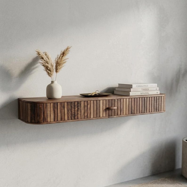 May include: A floating wooden shelf with a drawer and a curved front. The shelf is made of dark wood and has vertical grooves on the drawer front. A small white vase with dried plants, a tray with keys, and books are on the shelf.