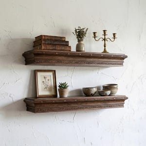 Könnte beinhalten: Zwei dunkelbraune Holzregale an einer weißen, strukturierten Wand. Die Regale sind mit Büchern, einem gerahmten Bild, einer kleinen Topfpflanze, Schalen, einer Vase mit getrockneten Blumen und einem Messing-Kerzenhalter dekoriert.