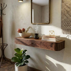 May include: A dark wood floating shelf with a drawer, displaying a bowl of flowers, books, and a decorative item. A mirror is above the shelf. A plant in a pot is below. The shelf has vertical grooves and is likely made of solid wood.