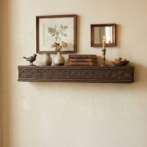 Puede incluir: Una estantería de madera tallada de color marrón oscuro con motivos florales. Muestra una impresión botánica enmarcada, un pequeño espejo, un candelabro de latón con una vela encendida, libros, jarrones con flores, un cuenco de madera y una figura de pájaro.