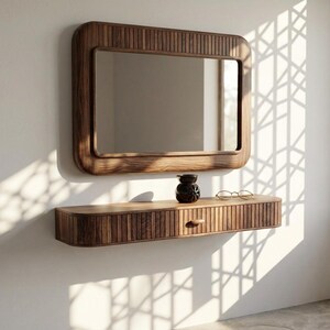 May include: A rectangular wooden mirror with a rounded frame is mounted above a matching floating shelf. The shelf has a single drawer and is adorned with a small vase and eyeglasses. The wood has a warm, natural tone.