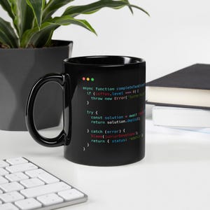 Puede incluir: Una taza de café negra con asa, con fragmentos de código coloridos. El texto incluye términos de programación como 'async function', 'try' y 'catch'. La taza está sobre una superficie blanca, con un teclado y una planta en el fondo.