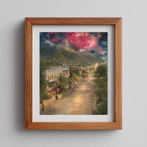 Op de afbeelding: Een ingelijste muurkunstprint met een stadsstraat bij nacht, met lichtsnoeren boven de weg, gebouwen aan de zijkanten en grote bergen op de achtergrond onder een donkere lucht vol met levendige rode en blauwe vuurwerkshows.