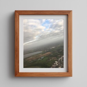 May include: A framed aerial view photograph showing a landscape with a lake, green fields, and buildings beneath a cloudy sky. The frame is light brown wood, and the photograph is set against a white mount.