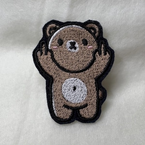 Può includere: Toppa ricamata raffigurante un orsetto dei cartoni animati con corpo marrone, contorno nero e pancia bianca. L'orso fa il dito medio con entrambe le mani e ha le guance rosa. La toppa è progettata per l'applicazione su abbigliamento o accessori.