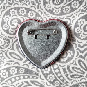May include: A heart-shaped pinback button with a metallic silver finish on the reverse side. The back features a horizontal steel safety pin mechanism securely attached to the center of the heart, designed for fastening the accessory to fabric.