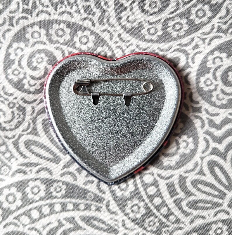 May include: A heart-shaped pinback button with a metallic silver finish on the reverse side. The back features a horizontal steel safety pin mechanism securely attached to the center of the heart, designed for fastening the accessory to fabric.