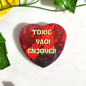 May include: A heart-shaped pin with a glossy finish featuring a dark red and black marbled background with iridescent glitter accents. The text TOXIC YAOI ENJOYER is printed in a bold, light yellow, rounded font across the center of the heart.