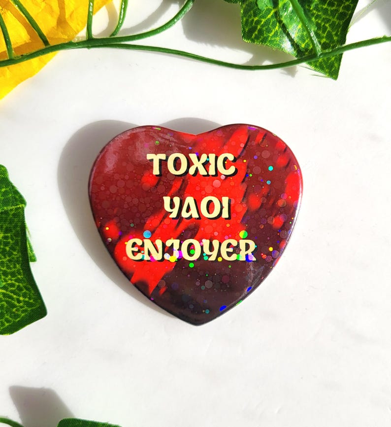 May include: A heart-shaped pin with a glossy finish featuring a dark red and black marbled background with iridescent glitter accents. The text TOXIC YAOI ENJOYER is printed in a bold, light yellow, rounded font across the center of the heart.