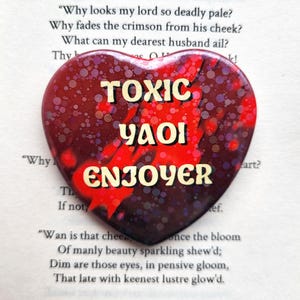 May include: A heart-shaped pin featuring a dark red and black background with a speckled, glittery texture. The text TOXIC YAOI ENJOYER is printed in bold, cream-colored capital letters across the center of the heart-shaped surface.