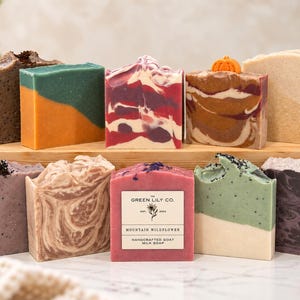 May include: A collection of ten rectangular bars of handcrafted goat milk soap in various colors and patterns, including one labeled The Green Lily Co. Mountain Wildflower, arranged on a wooden stand with a small orange pumpkin topper.