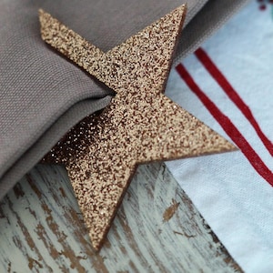 May include: A gold glitter star-shaped napkin ring adorns a folded gray napkin. The star is made of a textured material and adds a touch of sparkle. The napkin rests on a distressed wooden surface, with a patterned fabric visible.